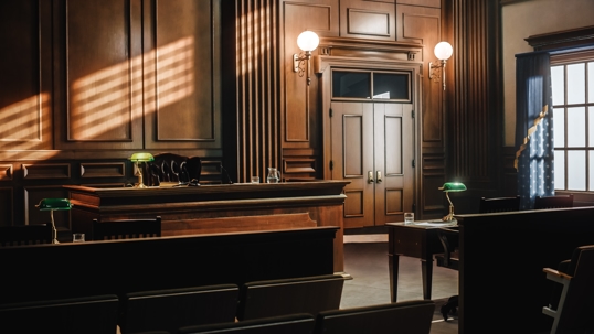 A well-lit, empty courtroom with a wooden judge's bench, green desk lamps, and rows of seats, bathed in natural light from the windows.